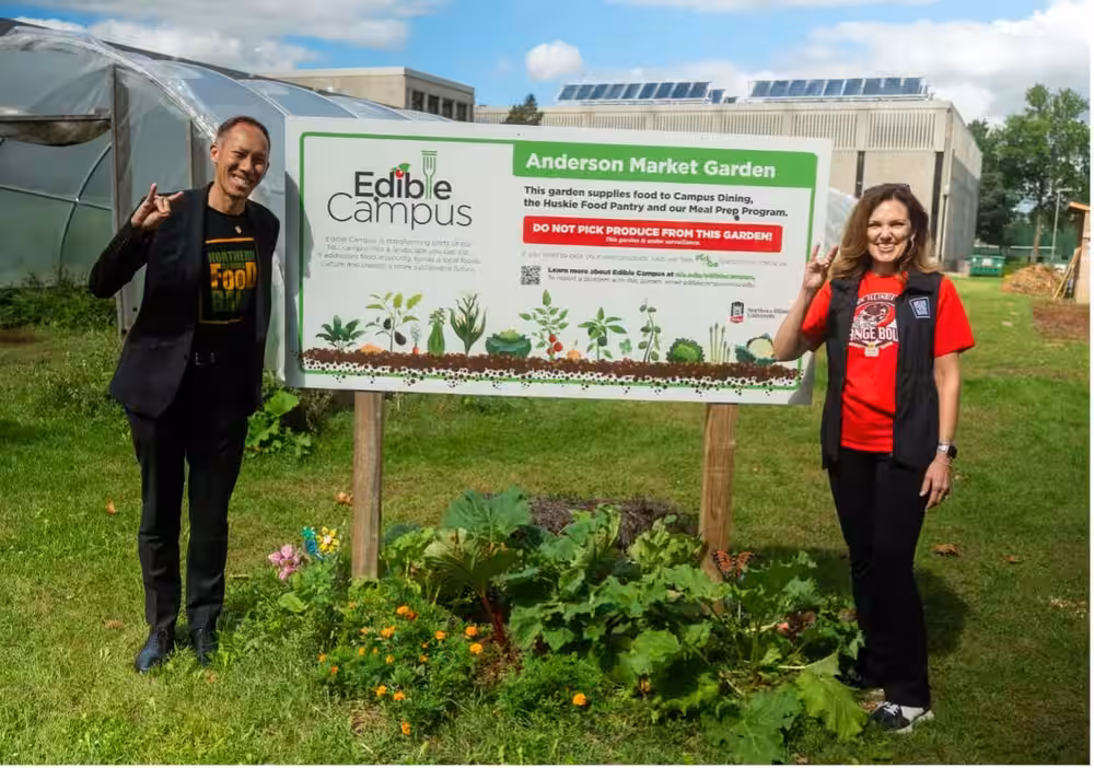 Joe Intrakamhang and Julie Yurko making the Huskie hand sign on either side of the Edible Campus sign at the Anderson Market Garden